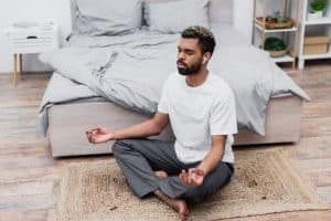 photo of man in lotus position on the floor next to a bed