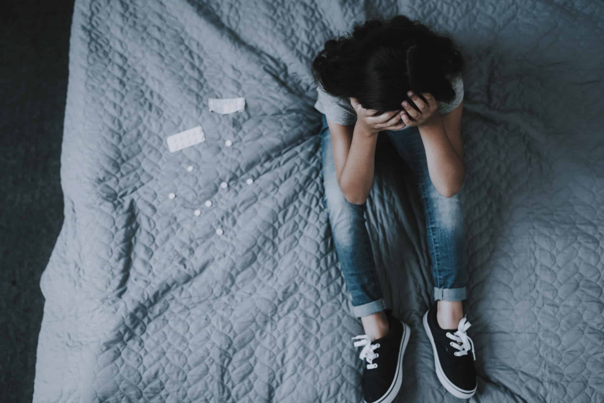 A teen girl sits on her bed with her head in her hands, and a few pills on the bed beside her, showing the frustration of running out of ADHD medications during the stimulant shortage.